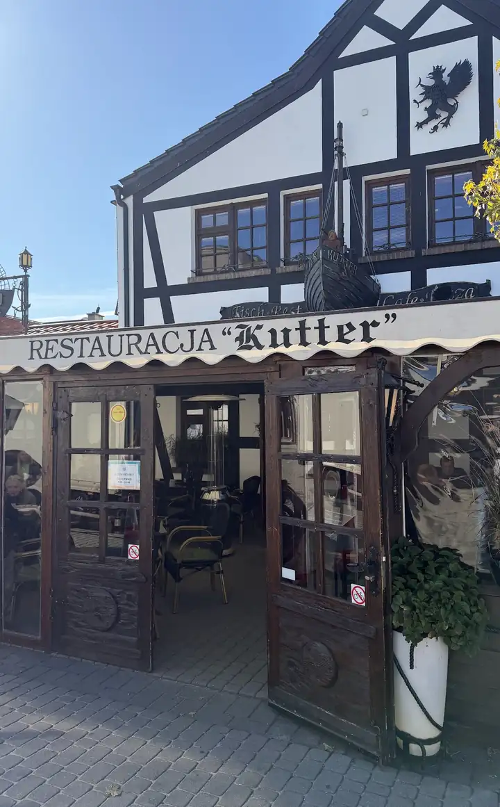 Biało-czarna fasada w stylu muru pruskiego z szyldem Restauracja Kutter i otwartymi drzwiami